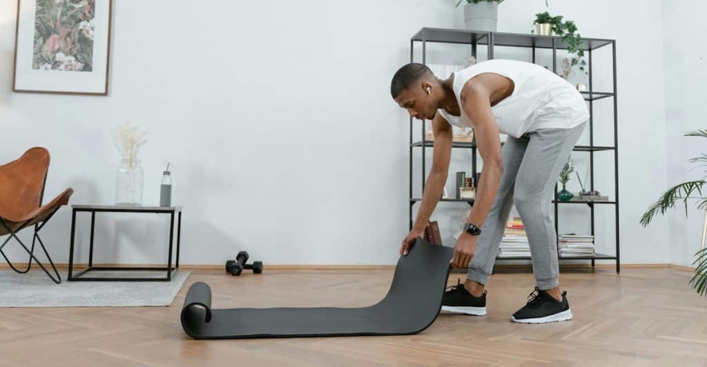 A man in a white tank top and gray sweatpants rolling up a grey yoga mat in a room with plants and furniture.