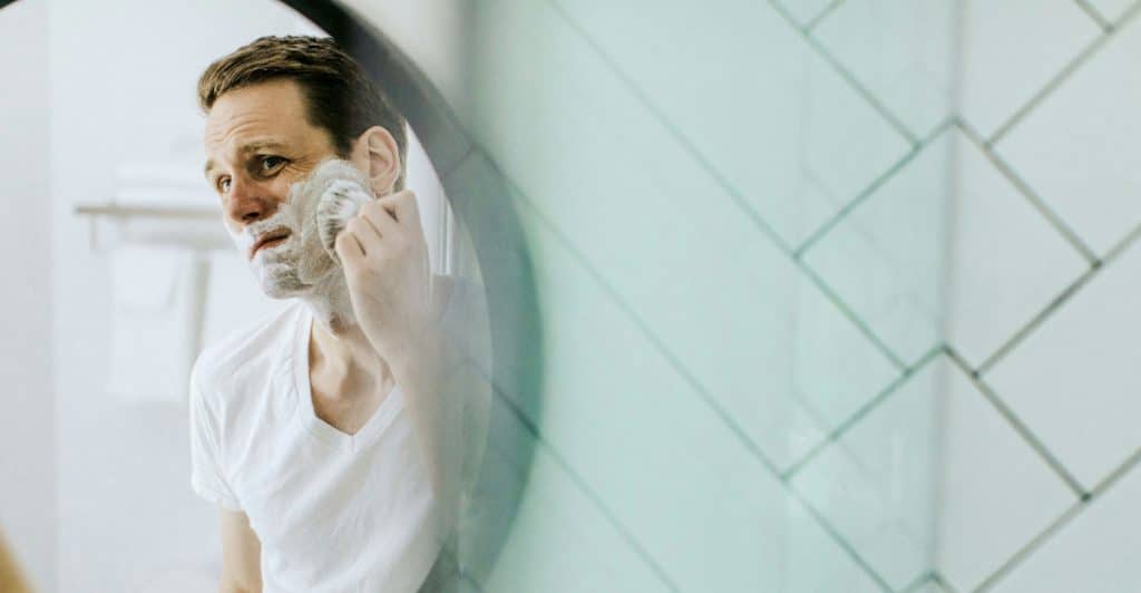 A man in a white t-shirt looking in a mirror applying shaving cream to his face in a tiled bathroom.