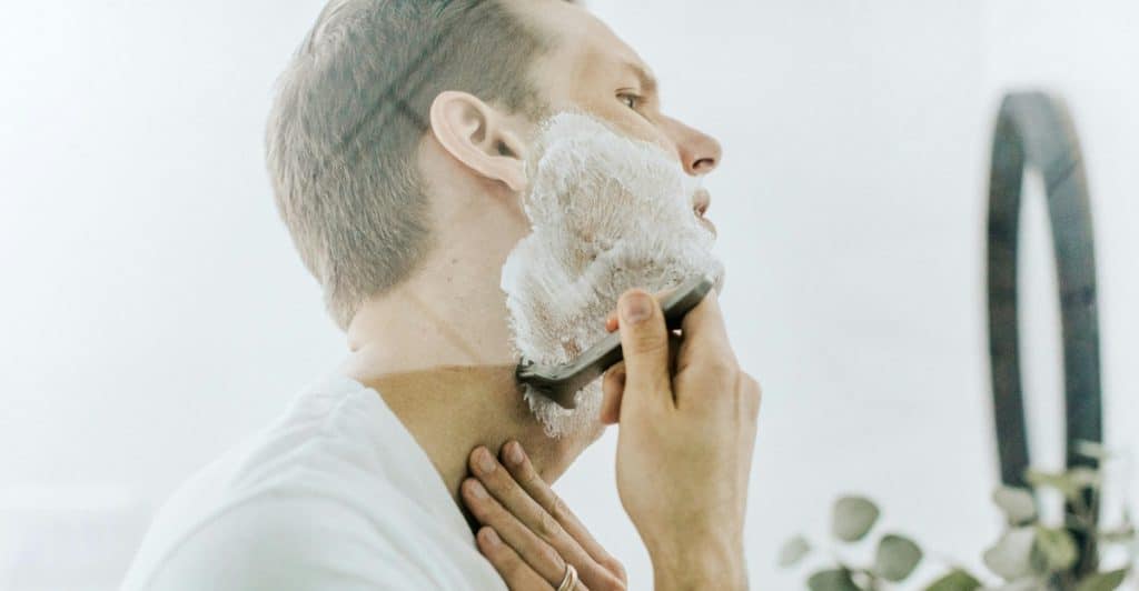 A man in a white t-shirt shaving his neck with a razor while looking in a bathroom mirror.
