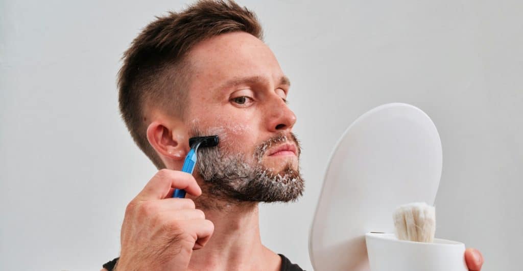 A man in a black tank top shaving his cheek with a disposable razor while holding a small white mirror.