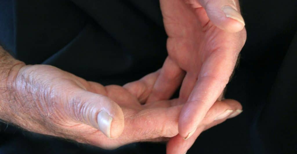 A close-up of two adult hands with neatly trimmed nails, clasped together against a dark, blurred background.