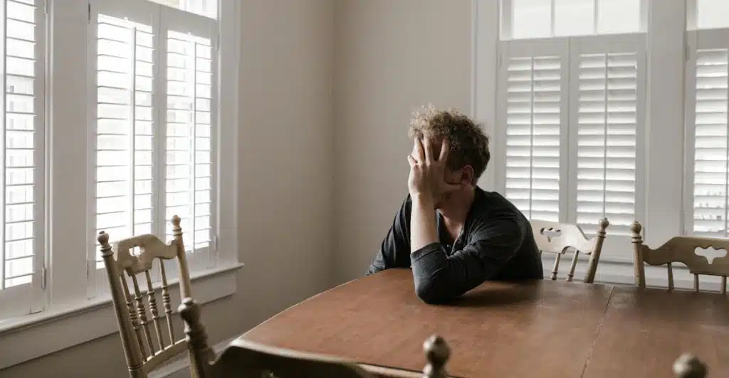 A man sits alone at a wooden table in a room with window shutters, covering his face with one hand, appearing stressed or contemplative.