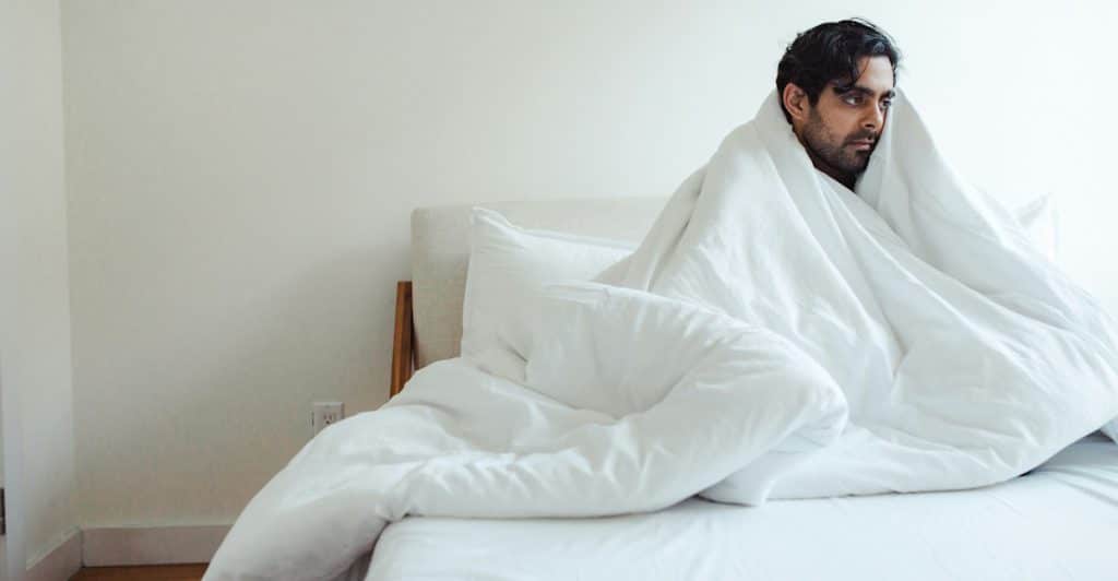 A man with dark hair who may be of South Asian descent sitting up in bed and wrapped in a thick white comforter.