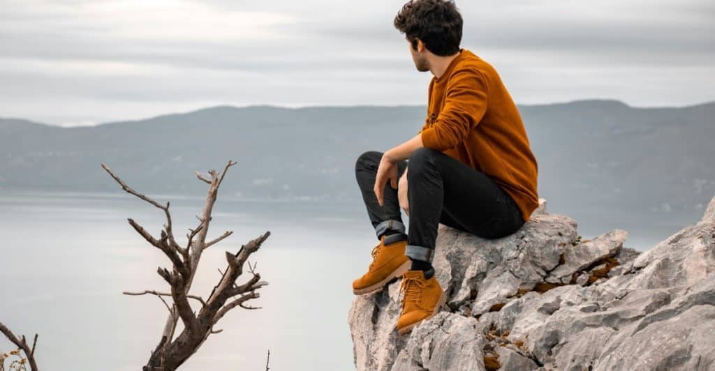 A man sitting on a rocky cliff overlooking a body of water with mountains in the distance.