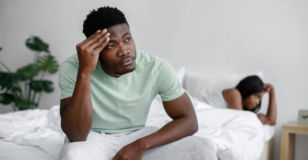 A man sitting on the edge of a bed, holding his head, with a woman lying in the background.