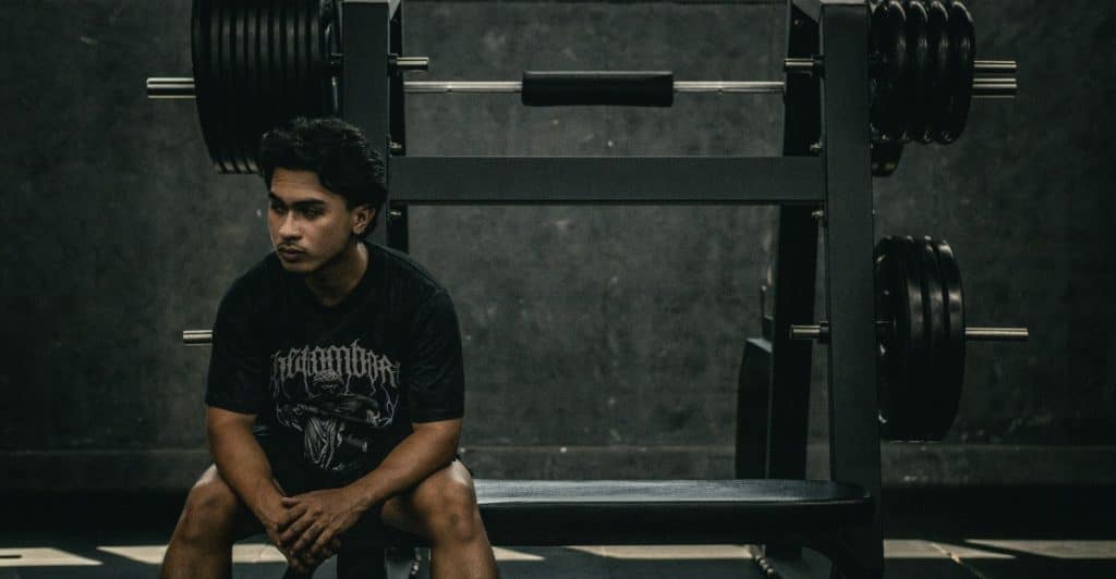A man in a black t-shirt and shorts is sitting on a gym bench, looking off to the side with a pensive expression, with weight racks visible behind him.
