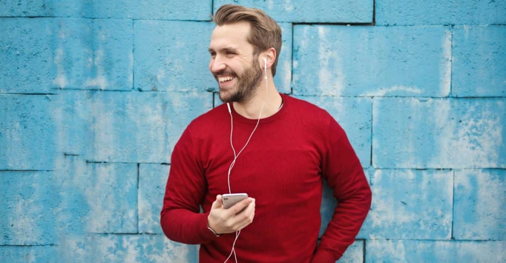 A man smiling and holding a phone, wearing headphones, against a blue textured wall.