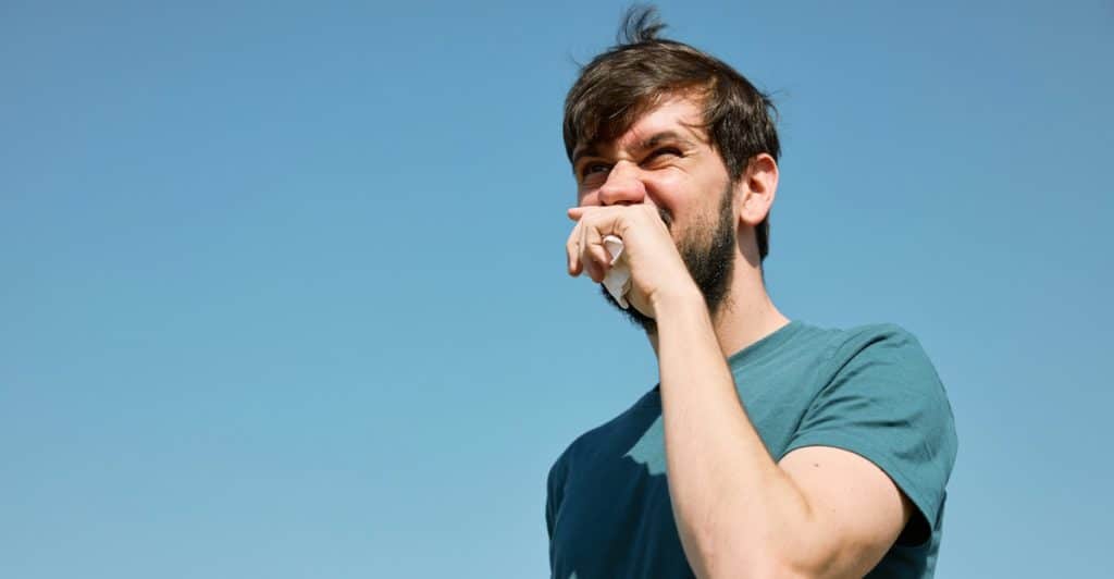 A bearded man in a teal t-shirt sneezing into a tissue against a clear blue sky.