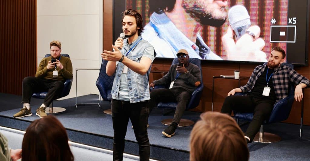 A man with a beard and a light-colored denim jacket is standing on a stage speaking into a microphone with his left hand extended, while several other men are seated in chairs on the stage behind him and a large screen.