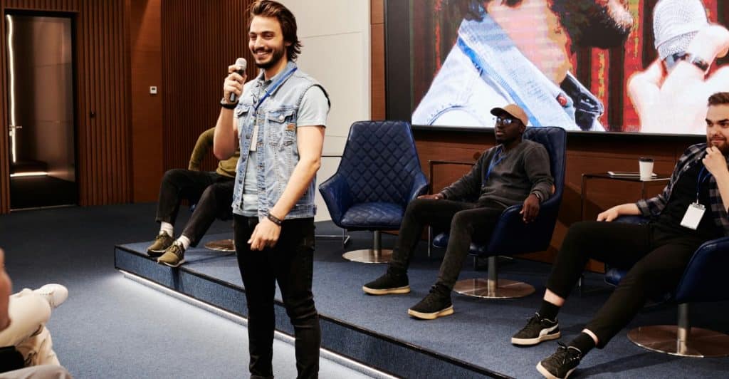 A man with long hair and a denim vest speaking into a microphone, standing in front of an audience and a large screen displaying an image.