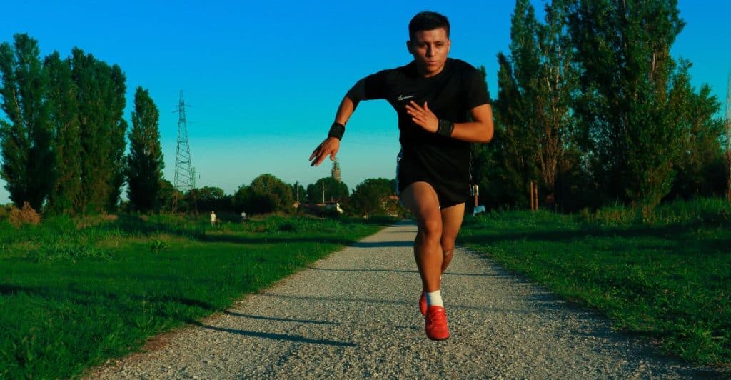 A young man in black shorts and a black t-shirt is sprinting towards the camera on a gravel path, with green grass and tall trees under a bright blue sky.