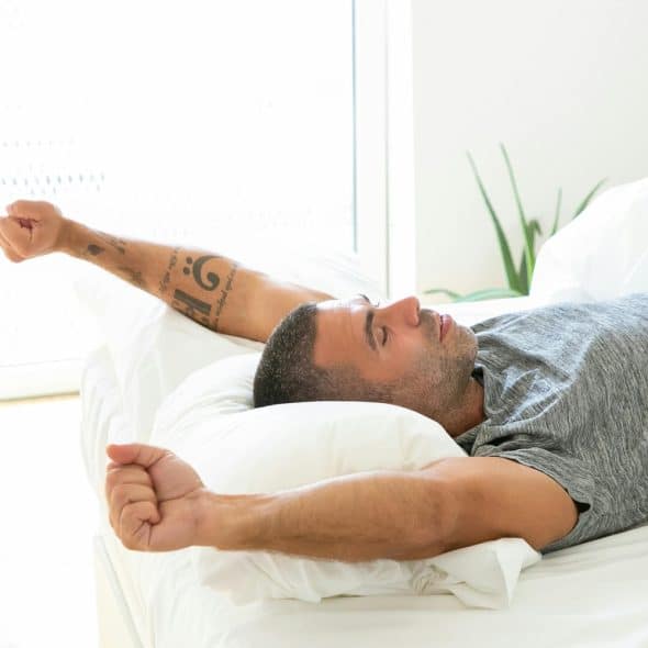 A man is stretching in a white bed with white sheets.