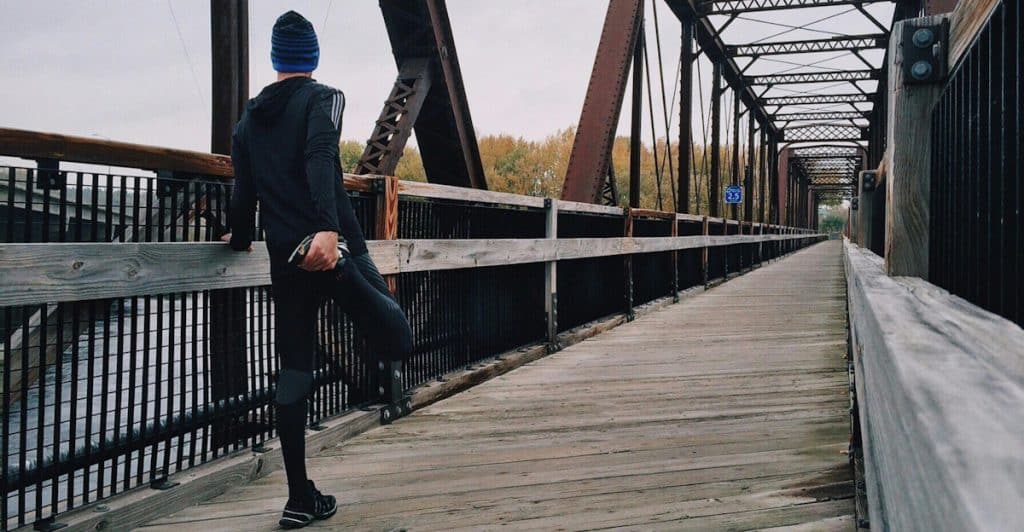 A man wearing athletic clothing and a beanie stretching on a wooden bridge next to a metal truss structure.