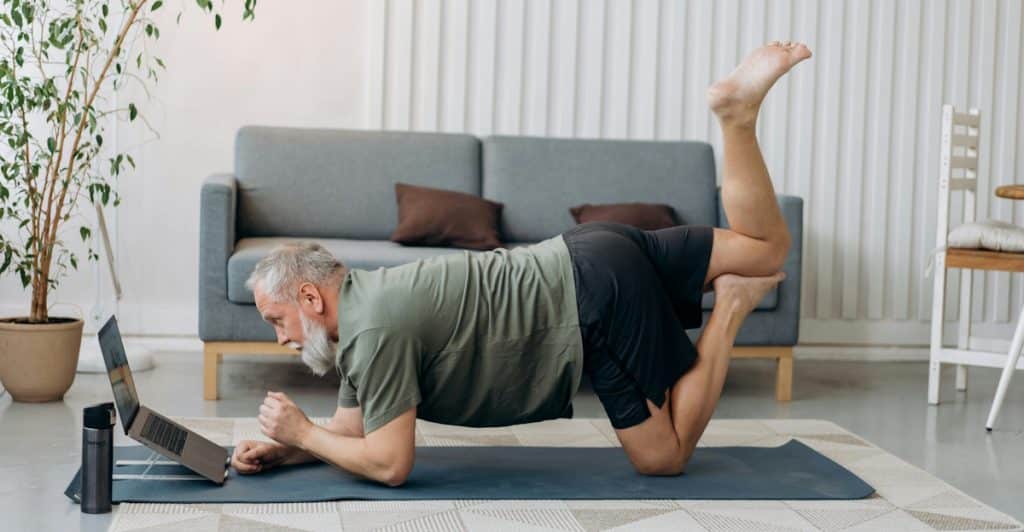 An older man with a beard is stretching on a yoga mat in a plank position, looking at a laptop in front of him.