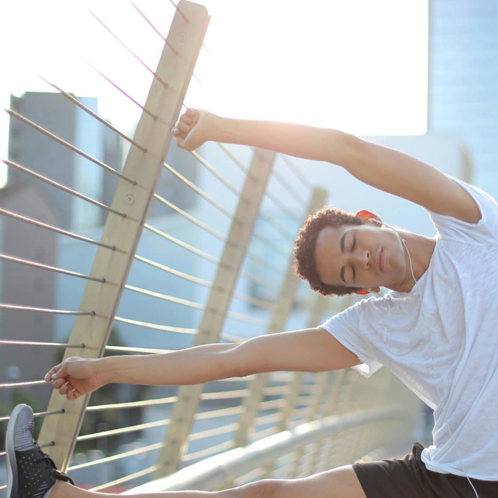 A man in a white t-shirt and black shorts stretching his leg on a railing with city buildings in the background.