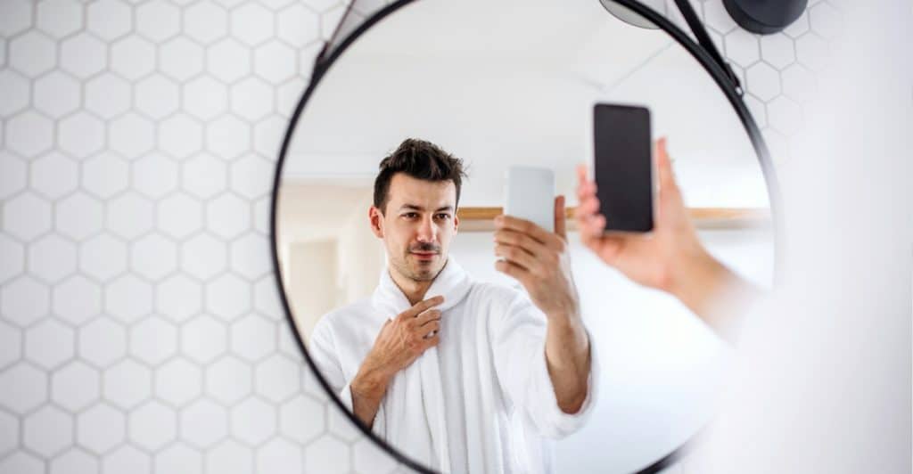 A man in a white robe is reflected in a round mirror hanging on a white hexagonal-tiled wall, as he holds up a smartphone to take a selfie.