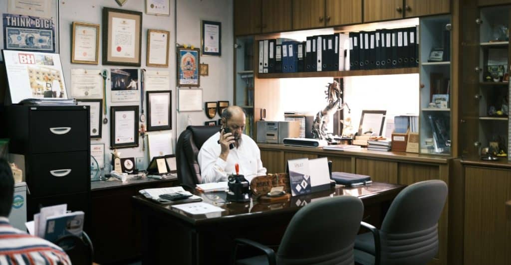 A man in a white shirt talking on a landline phone while sitting at a large wooden desk in an office filled with documents and shelves