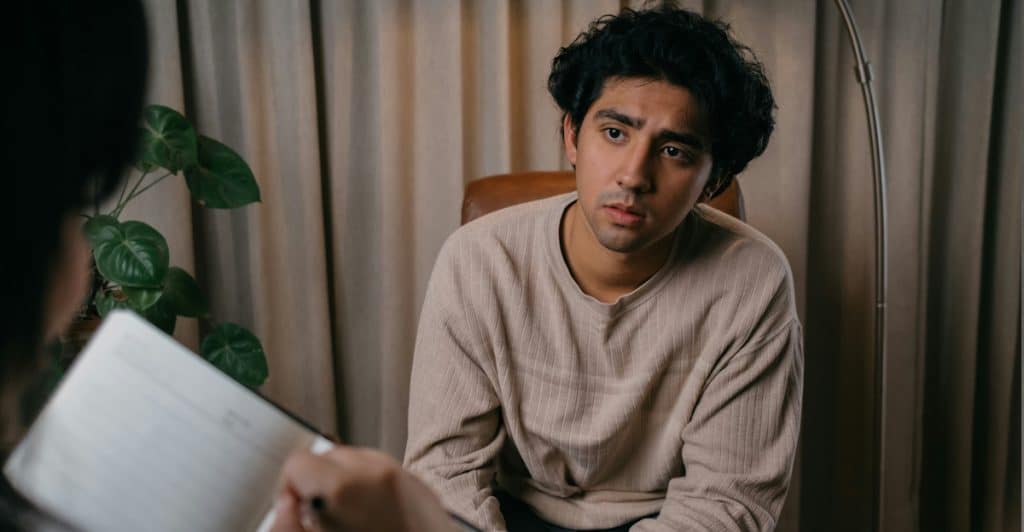 A young man sits in a chair, looking intently at someone off-camera who is holding a notebook, suggesting a therapy session.