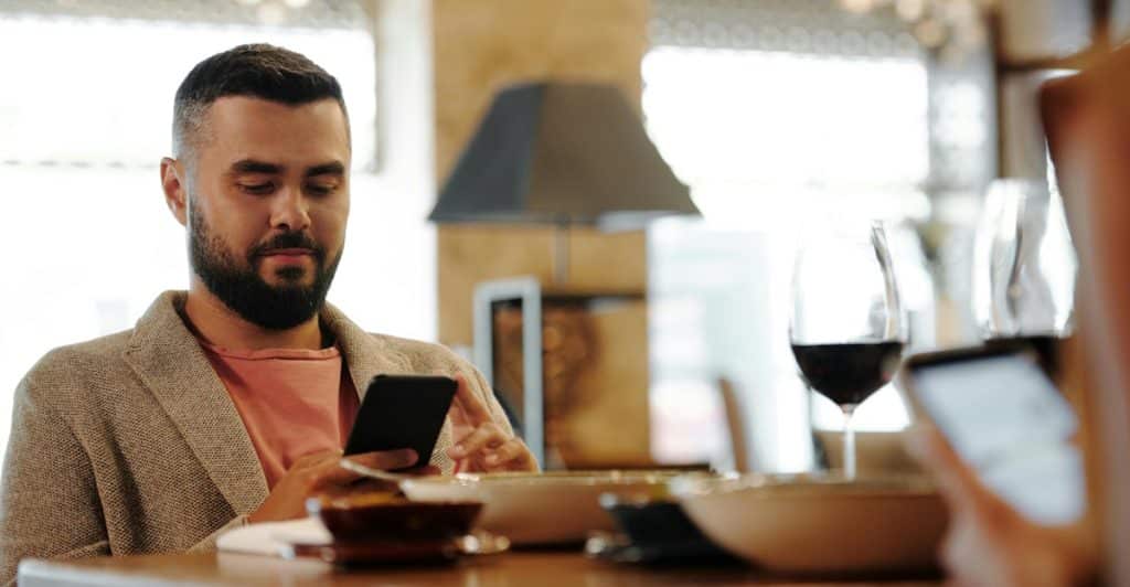 A man with a beard looking down at his phone while sitting at a table with a drink, with another person's hand also on a phone in the foreground.