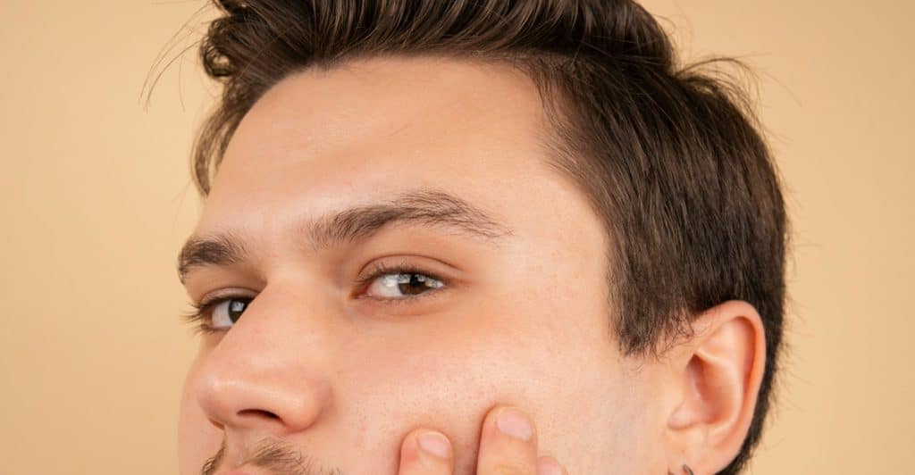 A close-up of a man's face with short facial hair, wearing two silver hoop earrings, as he touches his cheek with his right hand.