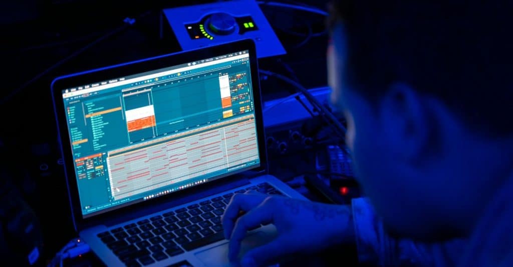 A man looking intently at a laptop screen displaying what appears to be music production software in a dark room.