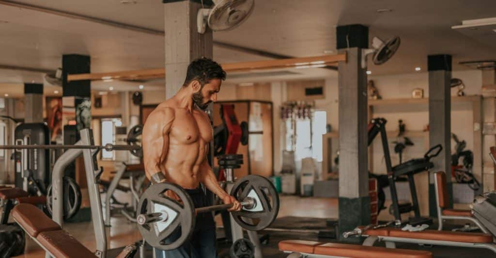 A shirtless, muscular man with a beard is lifting a barbell in a gym, with various gym equipment visible in the background.