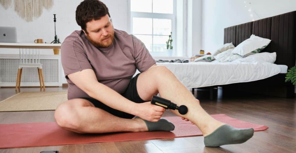 A man with a beard is sitting cross-legged on a red yoga mat, using a black handheld massage gun on his right calf muscle in a bedroom setting.
