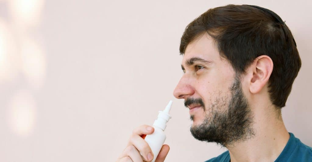 A profile view of a bearded man in a blue t-shirt using a white nasal spray bottle.
