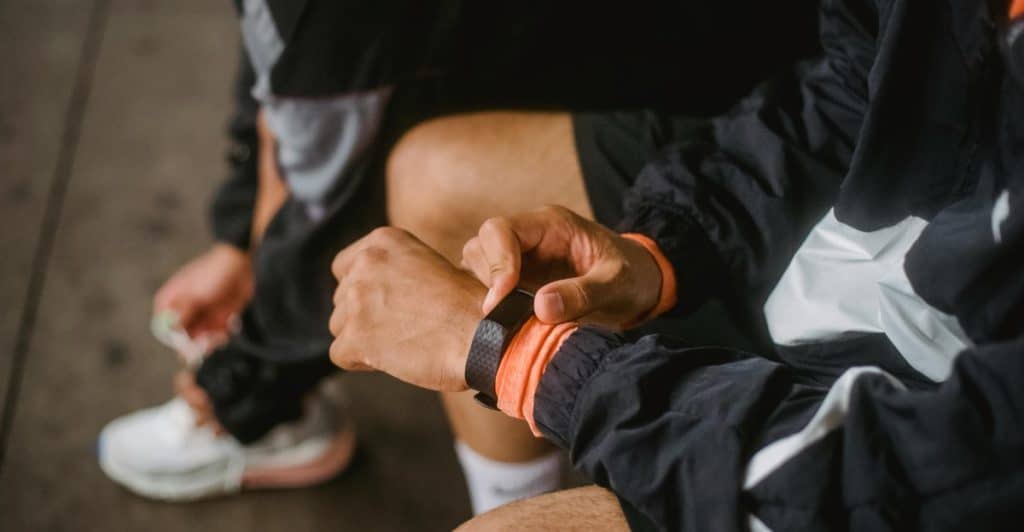 A person sitting down, adjusting a black and orange wristband on their arm, with another person's shoes visible in the background.