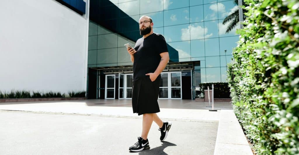 A man with a beard and glasses, dressed in a black t-shirt and black shorts, is walking while looking at his phone.
