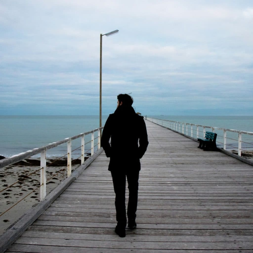 A man walking away from the camera on a long wooden pier over calm water with a cloudy sky.