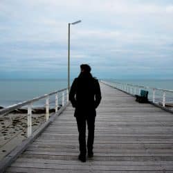 A man walking away from the camera on a long wooden pier over calm water with a cloudy sky.