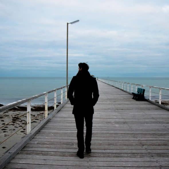A man walking away from the camera on a long wooden pier over calm water with a cloudy sky.