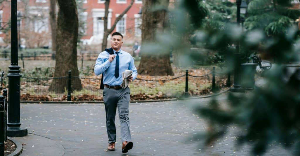 A man walking with good posture, holding a book and a jacket over his shoulder, in a park.