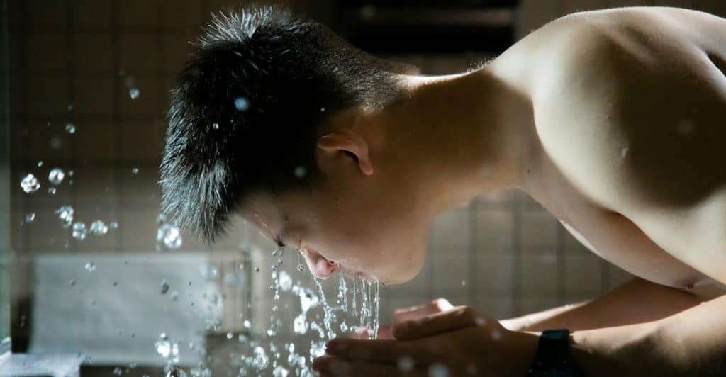 A shirtless man with dark hair splashing water on his face over a sink.