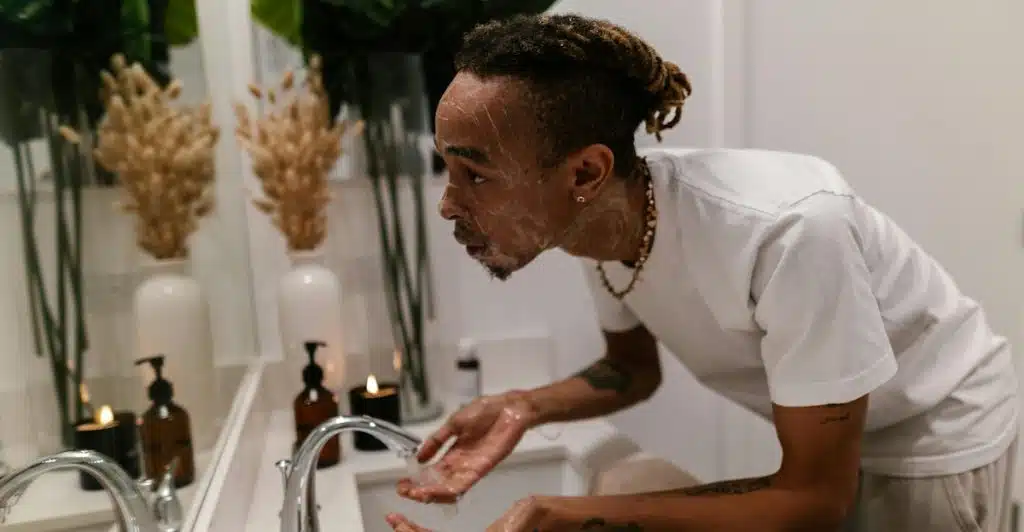 A man with dreadlocks is leaning over a bathroom sink, splashing water on his face, which has white foam on it.