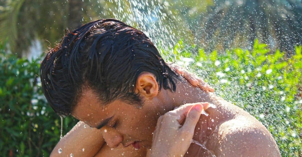 A shirtless man washing his hair under a shower outdoors.
