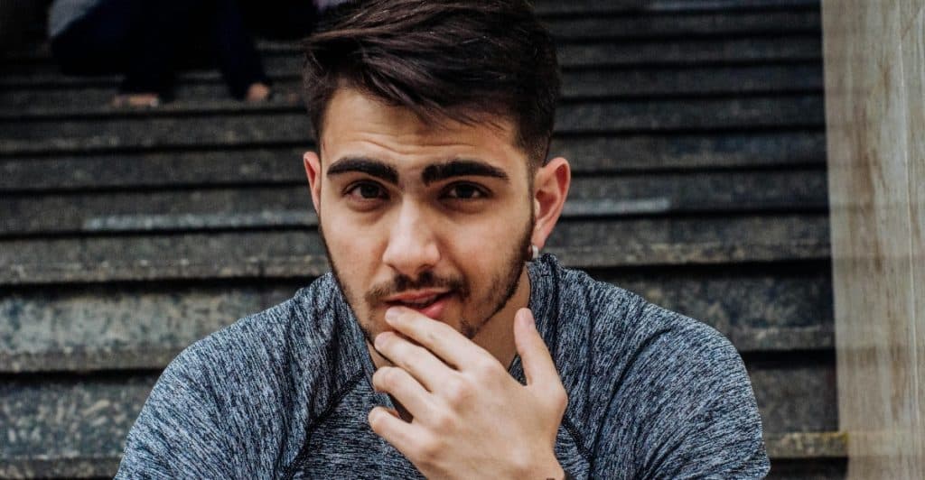 A man with dark hair and a beard wearing a grey speckled long-sleeve shirt and a black watch, looking towards the viewer.