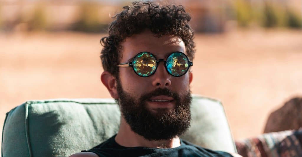 A man with a beard and curly hair is sitting in a green chair, wearing round sunglasses with colorful, kaleidoscopic lenses.