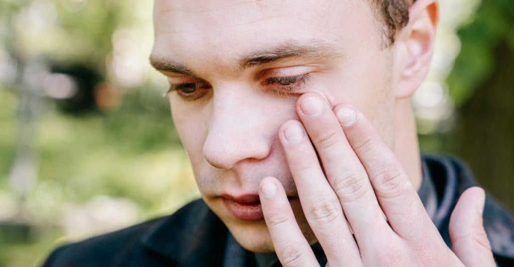A close-up of a man's face, with one hand near his eye, looking down with a sad or pensive expression.