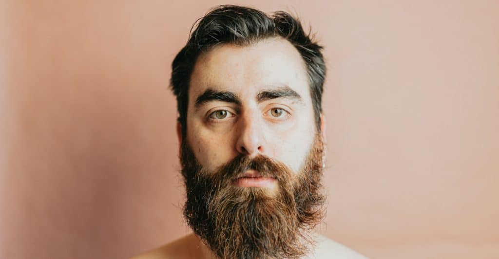 A shirtless man with a thick full beard and dark hair looking directly at the camera against a plain background.