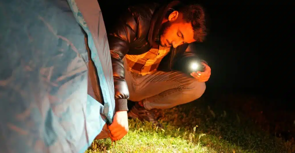 A man crouching outside a tent at night, illuminating the ground with a small, compact flashlight