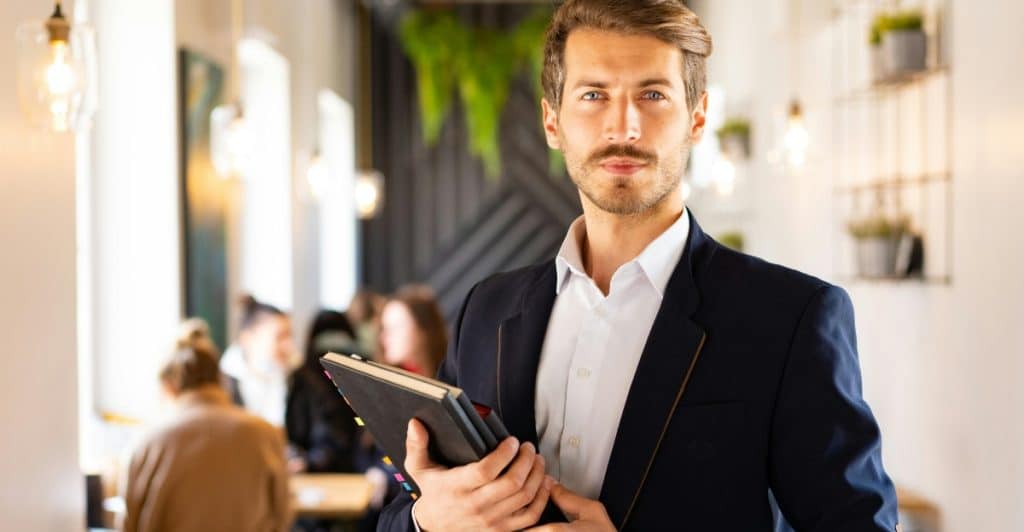  A man with a neatly trimmed mustache and stubble, wearing a suit jacket and white shirt, holding notebooks in an indoor setting.