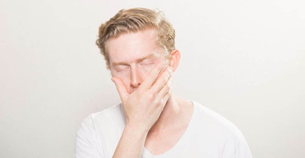 A man with reddish-blond hair and freckles in a white t-shirt with his eyes closed and hand covering his mouth and cheek.
