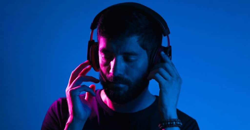 A man with a beard wearing black over-ear headphones, with his eyes closed and hands near the headphones, against a blue and pink gradient background.