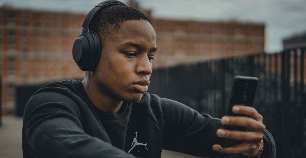 A man wearing black over-ear headphones and a dark long-sleeved shirt, sitting outdoors and looking intently at his smartphone.