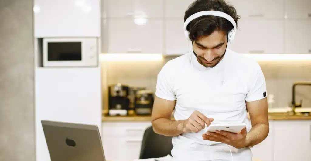 A man with headphones looking down at a tablet, with a laptop open on a table next to him in a kitchen setting.