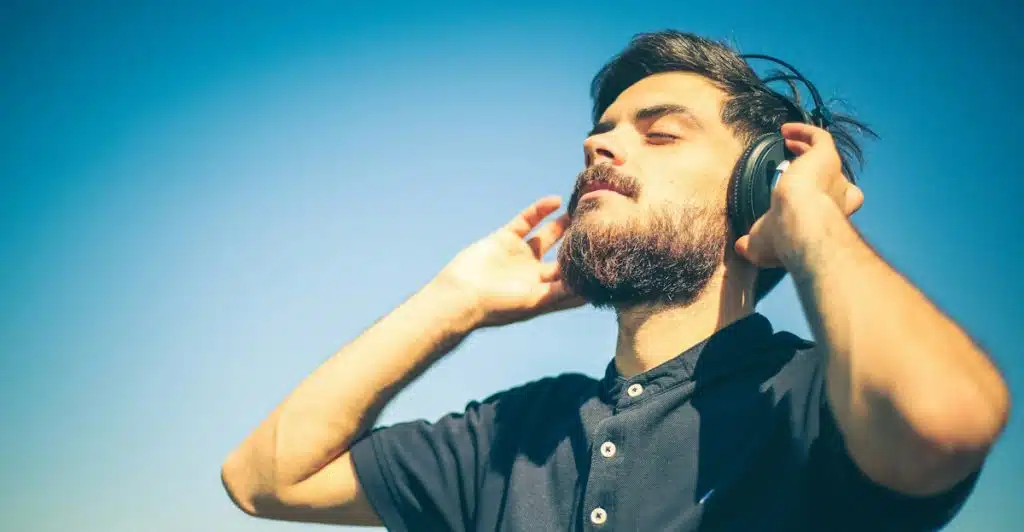 A bearded man wearing black over-ear headphones, with his head tilted back and eyes closed, standing outdoors under a clear blue sky.