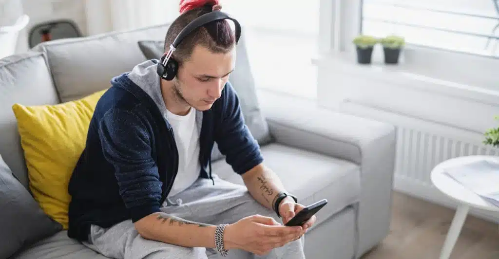 A man with red hair and headphones sitting on a grey couch, looking at and holding a smartphone.