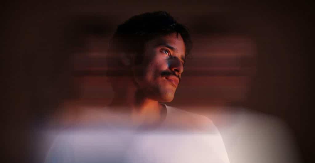 A man with a mustache and dark hair looks upwards and to the side, with a soft, glowing light effect around him.
